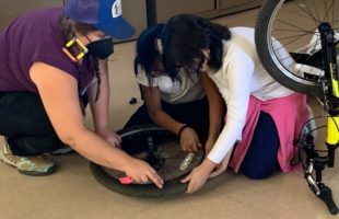 An adult teaches two youth how to repair a bicycle tire